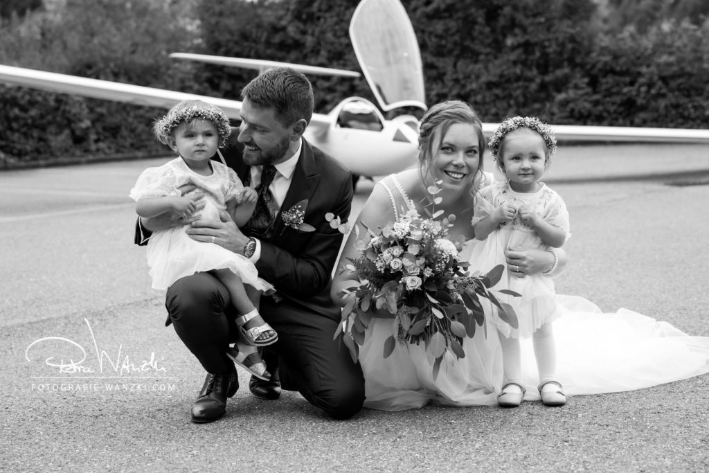 Brautpaar mit zwei Mädchen werden von Fotografin Winterthur vor einem Segelflieger kniend fotografiert, Bräutigam lacht Ringmädchen an, beide Mädchen haben Blumenkränze auf, die Braut hält einen modernen Brautstrauss