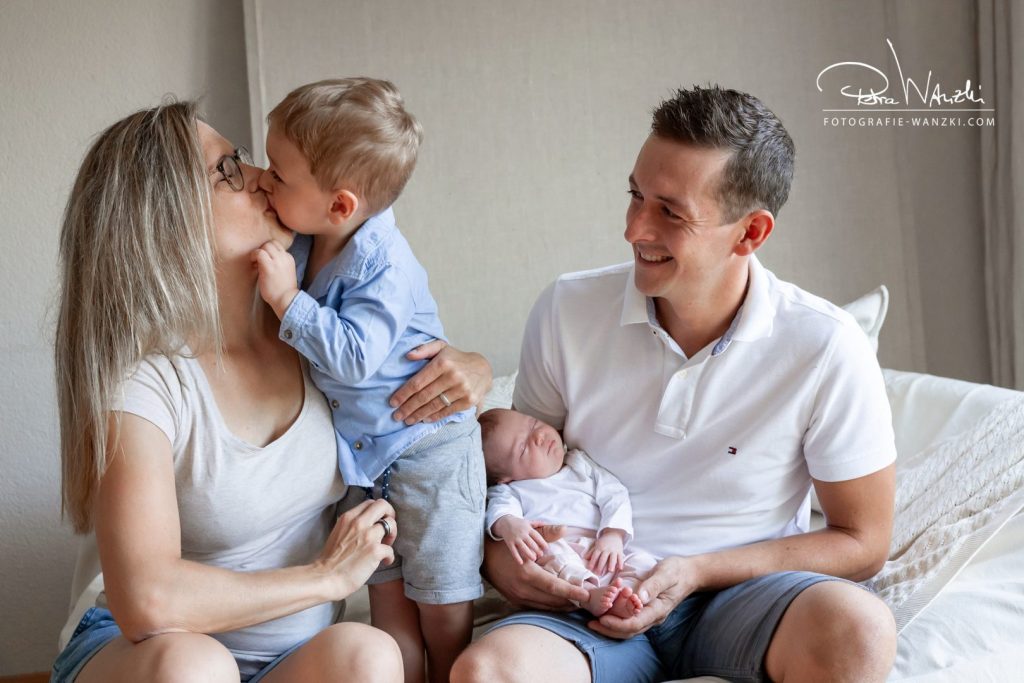 Familie mit Baby und Geschwisterkind, älteres Kind küsst Mama, Papa hält das Baby und freut sich sehr