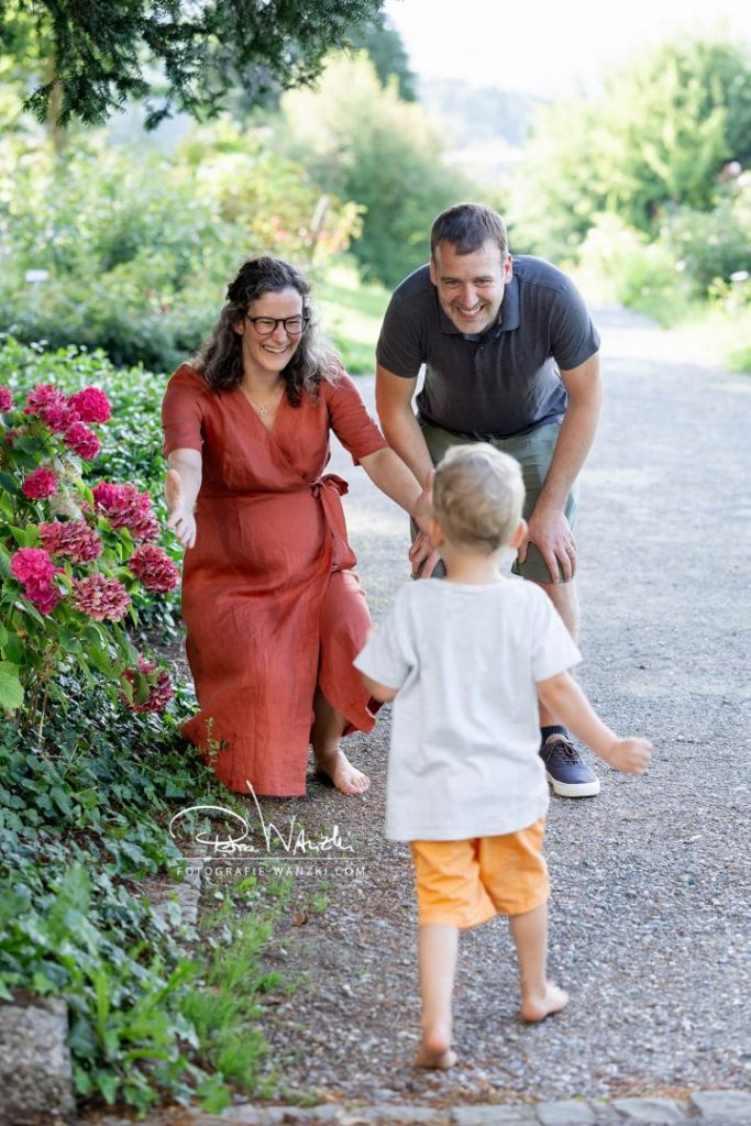 Eltern lachen, freuen sich auf das entgegenrennende Kind, Mutter ist Schwanger, im Park mit blühenden Blumen in Winterthur