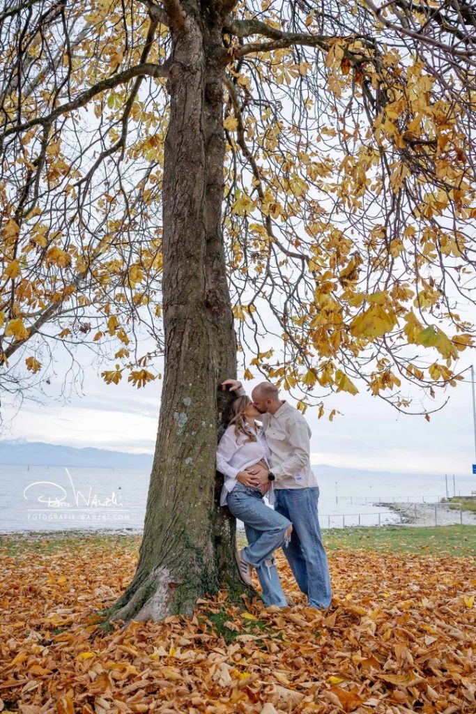 Schwangerschaftsfotoshooting im Herbst am See – werdende Eltern küssen sich liebevoll unter einem Baum mit gelben Blättern