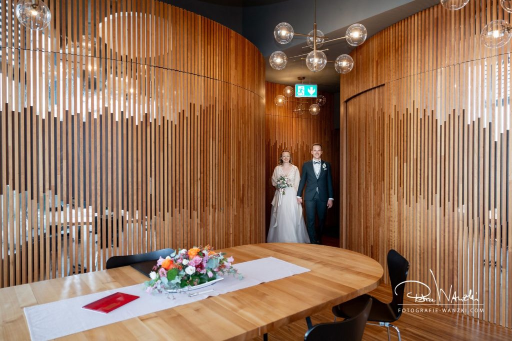 Brautpaar geht Hand in Hand in das Zivilstandesamt im Tower in Zug, der Raum ist mit moderner Holzvertäfelung, gerundet, moderne Lampen an der Decke und Blumenbouqet auf dem Tisch