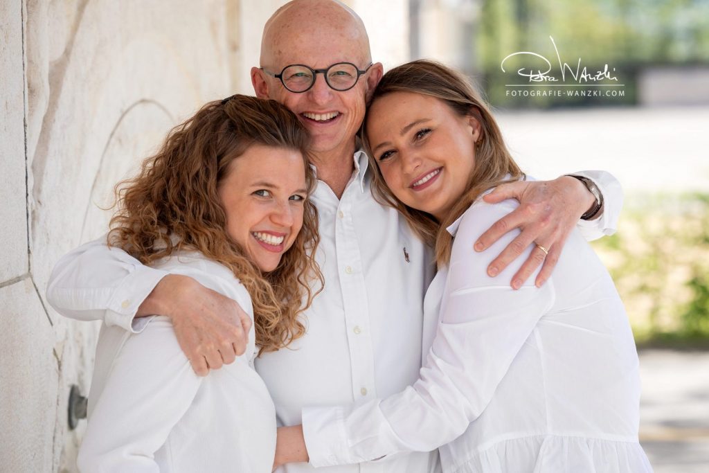 Vater umarmt seine Töchter liebevoll, alle in weiss gekleidet, offen, hell und glücklich