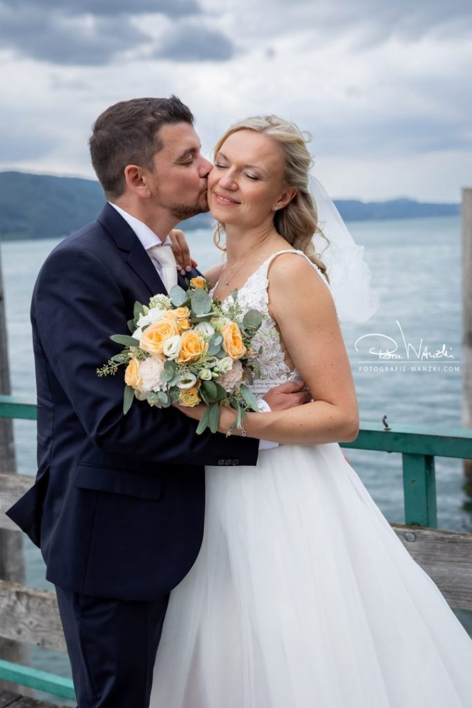 Bräutigam gibt Braut einen Kuss auf die Wange, See im Hintergrund, Braut hält Brautstrauss beim FotoShooting für die Hochzeitsbilder in der Hand