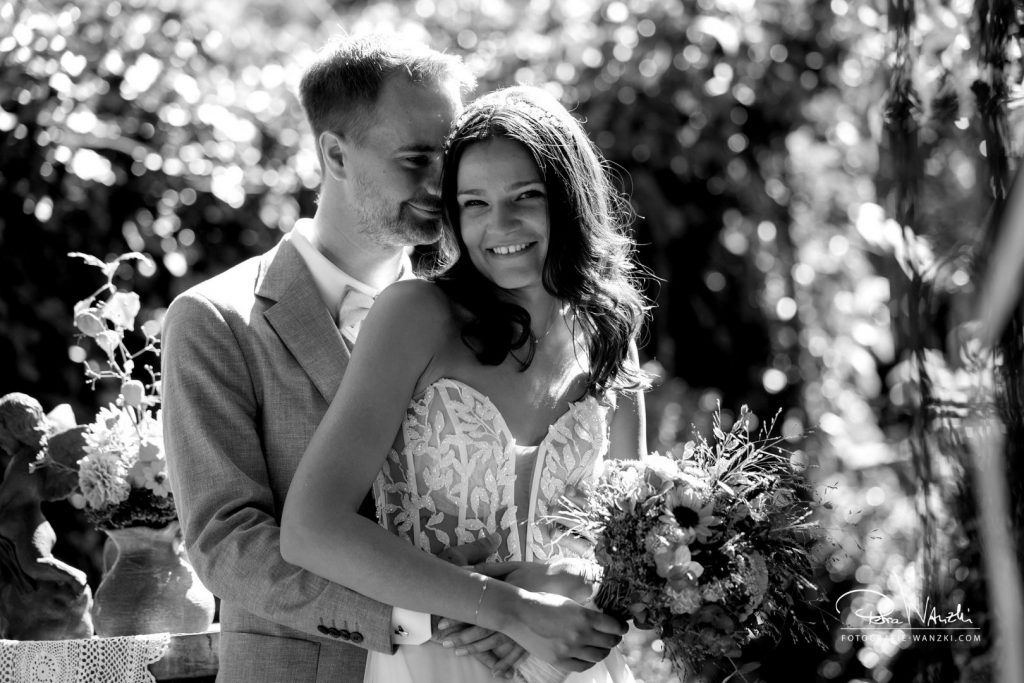 Bild in schwarz-weiss, Bräutigam Umart die Braut um die Taille und flüstert ihr etwas ins Ohr