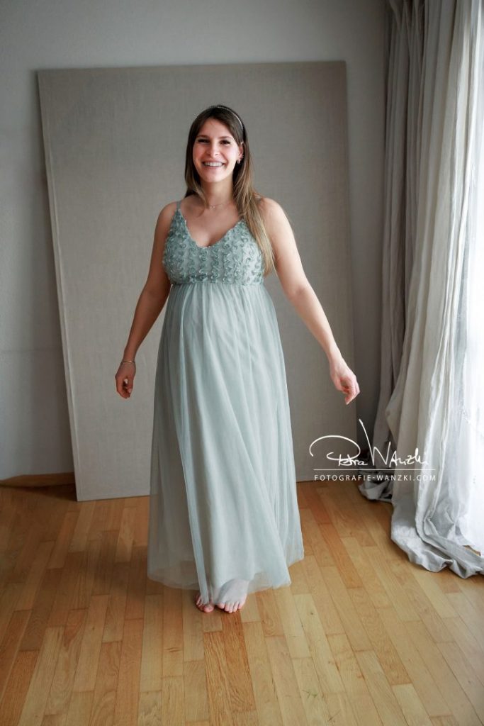 im Studio trägt eine Schwangere ein Eukalyptus farbenes Schwangerschaftskleid für FotoShooting in Winterthur, sie dreht sich und lacht, das Kleid schwingt, das Kleid ist aus Tüll mit Trägern