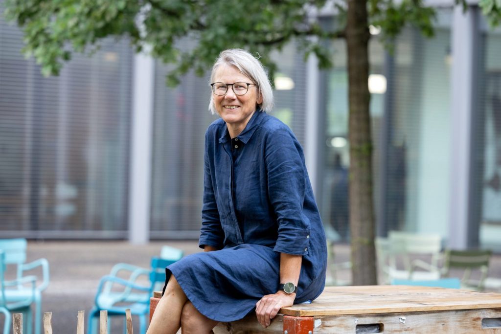 Frau in blauem Leinenkleid sitzt auf einer Palette im Park und lächelt in die Kamera, Bäume und türkisfarbene Stühle im Hintergrund sichtbar