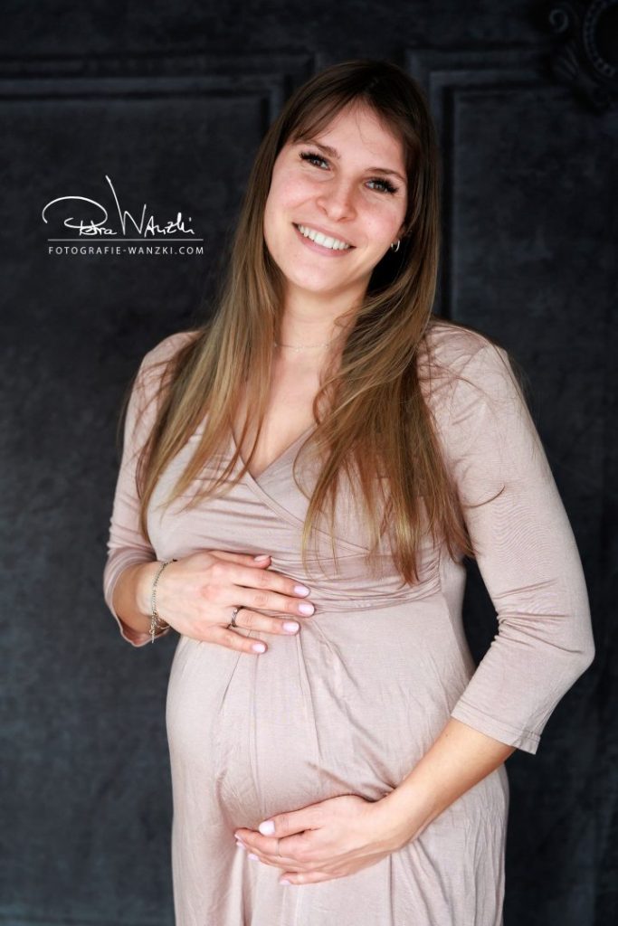 Schwangere Frau mit langen Haaren trägt ein rosa Jersey Kleid als Outfit und hält ihren Babybauch mit beiden Händen, vor schwarzem Hintergrund