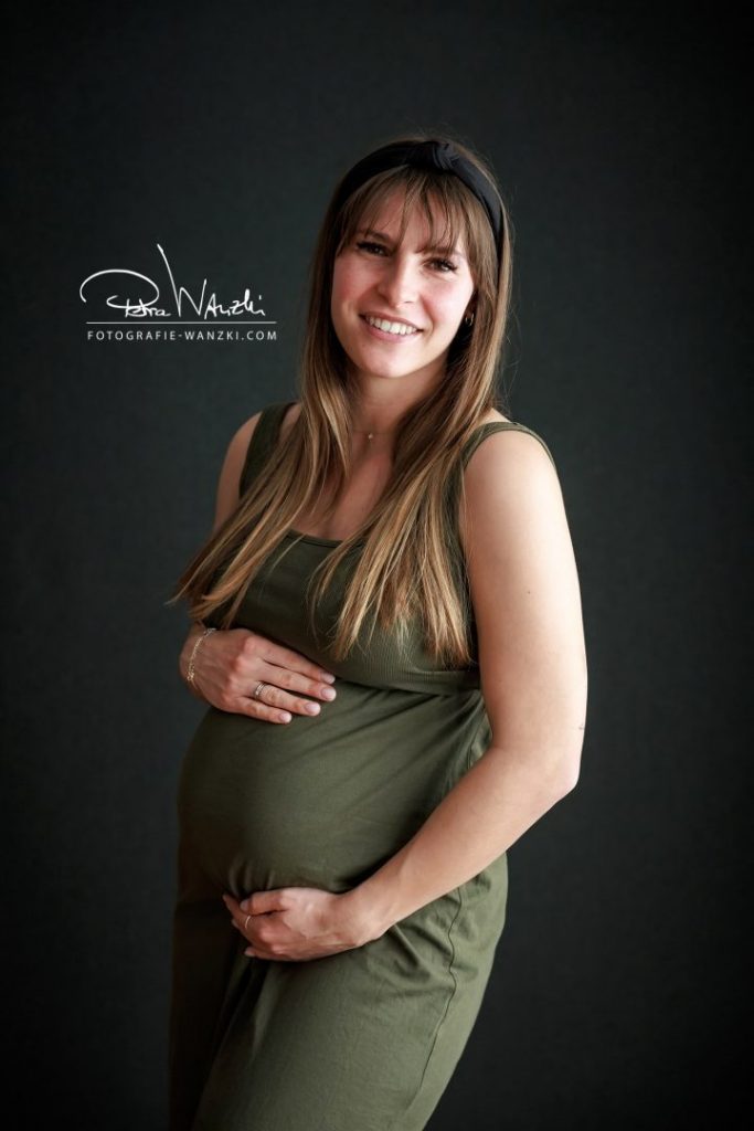 Schwangere mit BabyBauch im Outfit für das BabyBauch FotoShooting im Studio, sie trägt ein oliv grünes, eng anliegendes Trägerkleid aus Feinripp Stoff