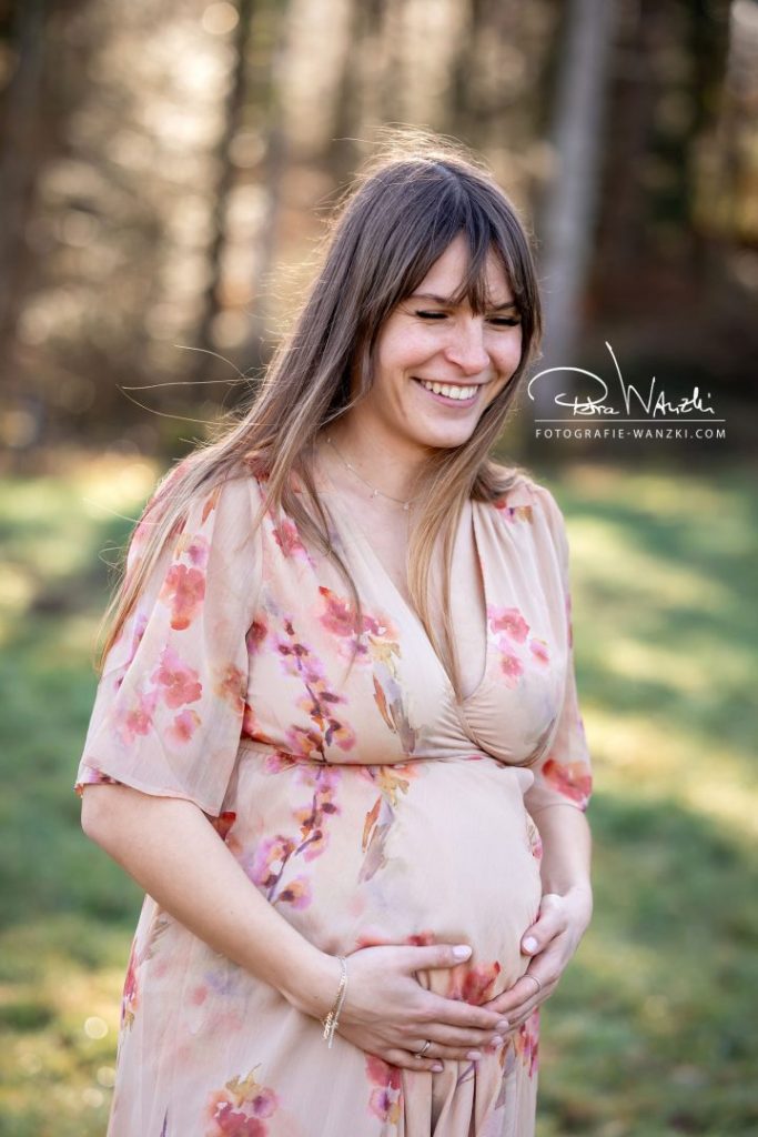 schwangere Frau lacht glücklich mit geschlossenen Augen, sie hält die Hände über den Babybauch, trägt ein rosa geblümtes Kleid
