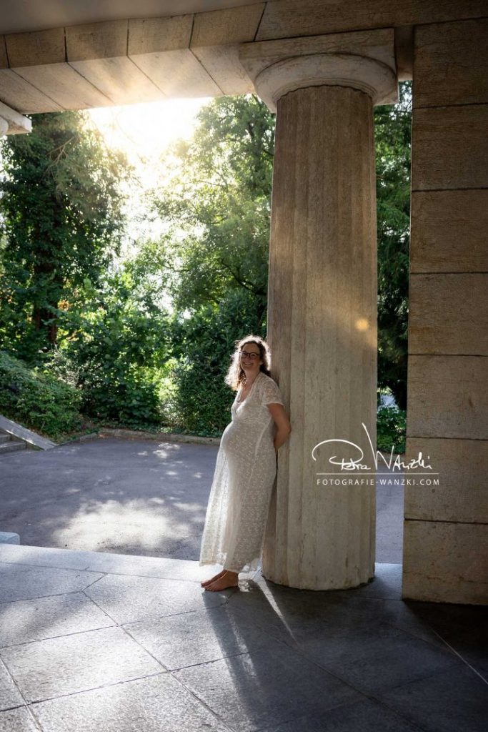 Beim Babybauch FotoShooting steht Frau mit Babybauch bequem an eine Säule gelehnt, die Abendsonne scheint von hinten durch die Bäume, wirkt lichtdurchflutet, riecht nach Sommer, Outfit ist ein helles Schwangerschaftskleid
