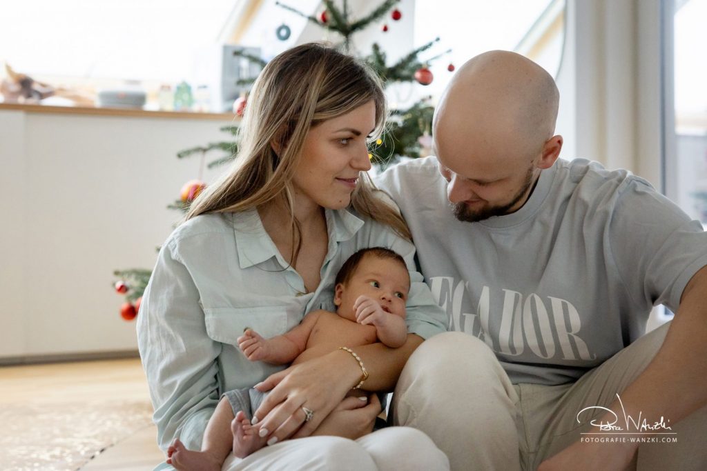 Familie sitzt beim FotoShooting in Zürich mit neugeborenem Baby auf dem Arm zuhause v or dem Weihnachtsbaum, Vater schaut auf das Baby, Mutter schaut verliebt den Papa an